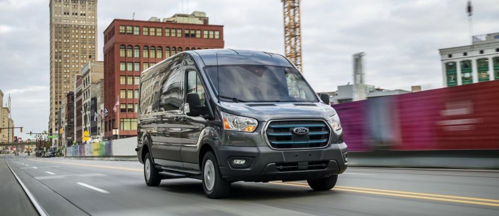 Een Ford E-Transit rijdt over een brede weg met flatgebouwen op de achtergrond