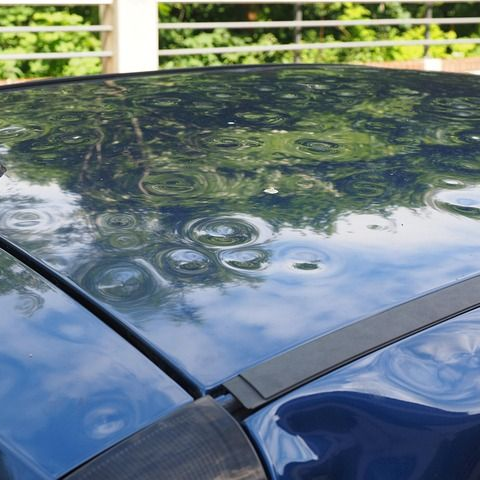 Blauw autodak beschadigd door hagel, de weerspiegeling van de groene bomen en het witte hek.