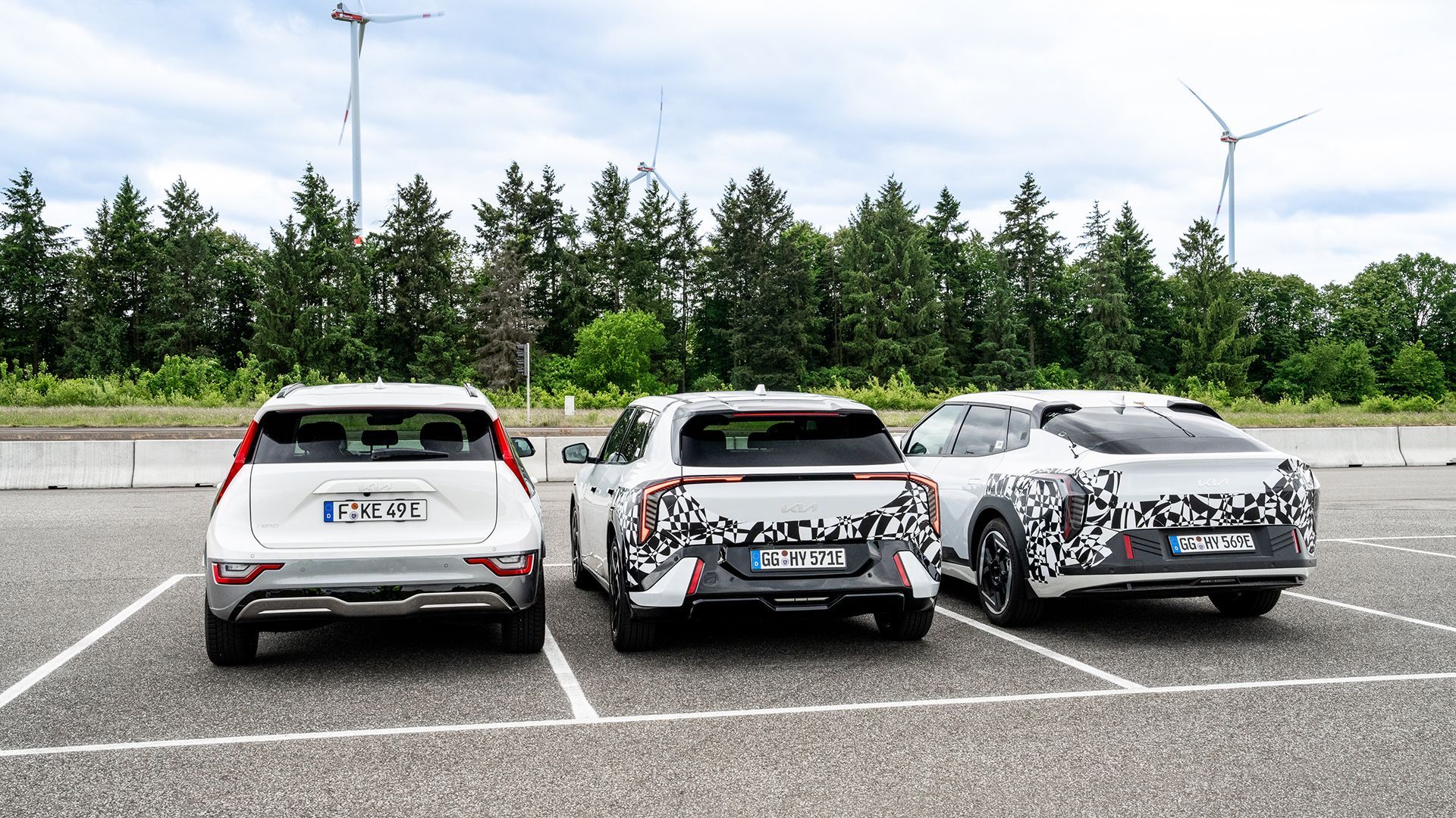 Three white KIA EV4 car parked in a lot, with the middle car covered in camouflage. Hatchback in the middle. EV4 Fastback on the right.