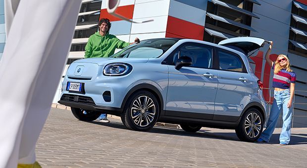 Een man en een vrouw staan op straat bij een lichtblauwe leapmotor T03 met de achterklep open