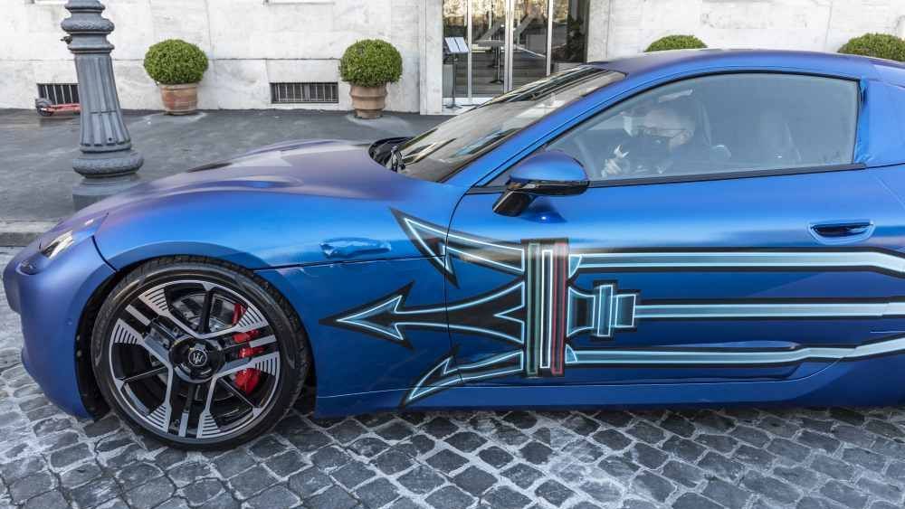Een blauwe Maserati GranTurismo staat voor een wit gebouw op een straat met klinkers