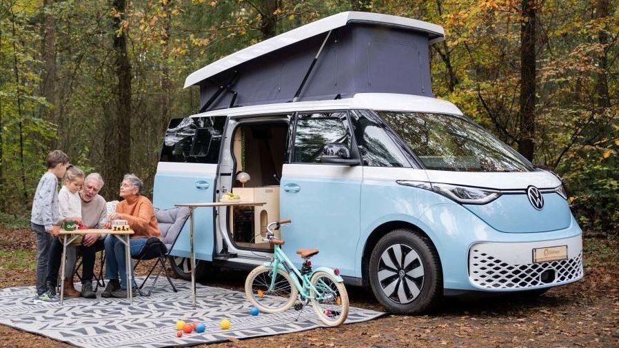 Een familie zit ergens in het bos aan tafel bij een elektrische campervan Outbase ID.Buzz