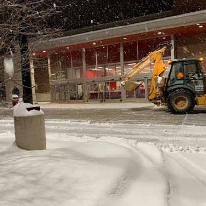 A yellow excavator is clearing snow from the sidewalk in front of a building.