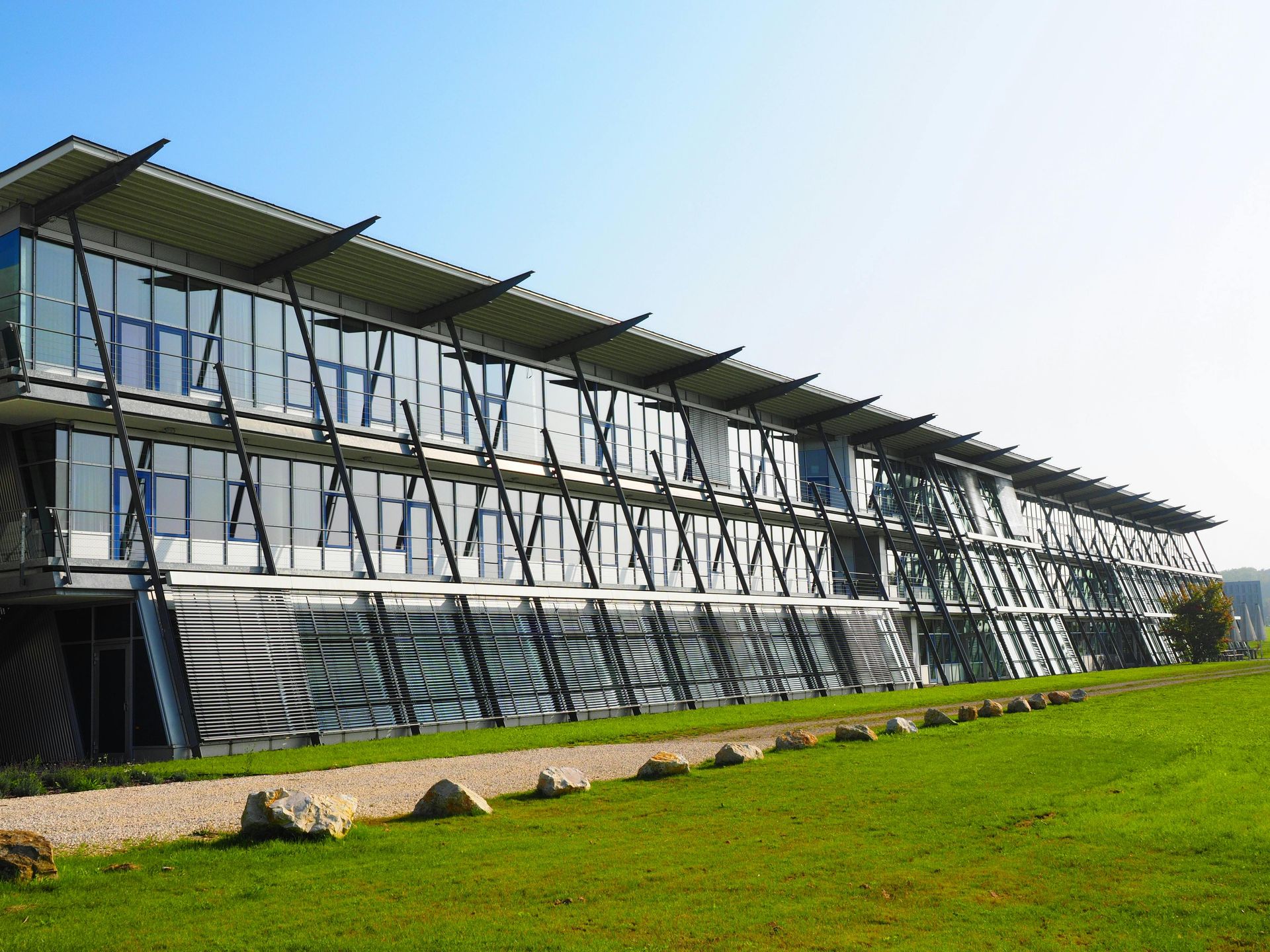Modern glass and steel building with angled supports, situated on a green lawn with a clear blue sky.