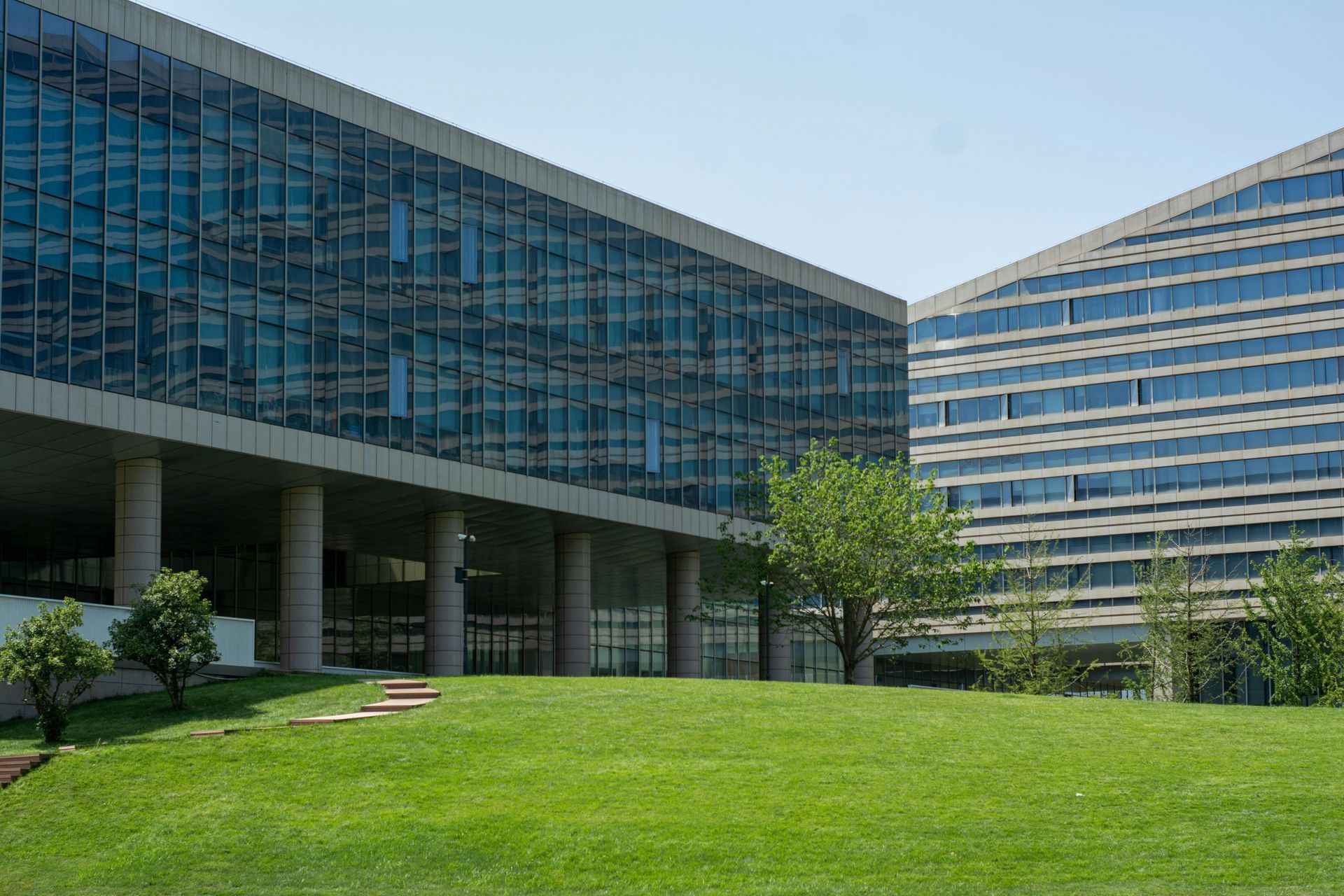 Two modern office buildings with glass facades stand behind a green lawn and small trees.