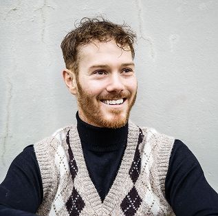 A man with a beard is wearing a sweater vest and smiling.