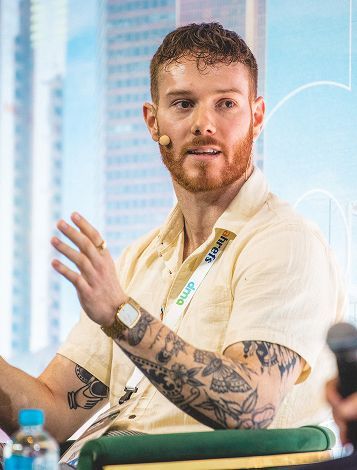 A man with a beard and tattoos is sitting in front of a microphone.H