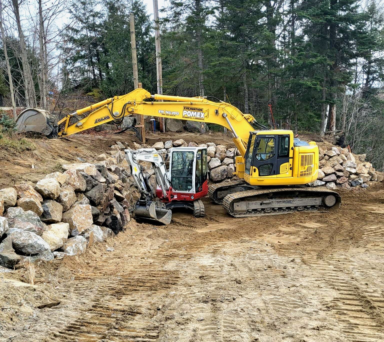 Deux excavatrices travaillent sur un mur de pierre dans un champ de terre.