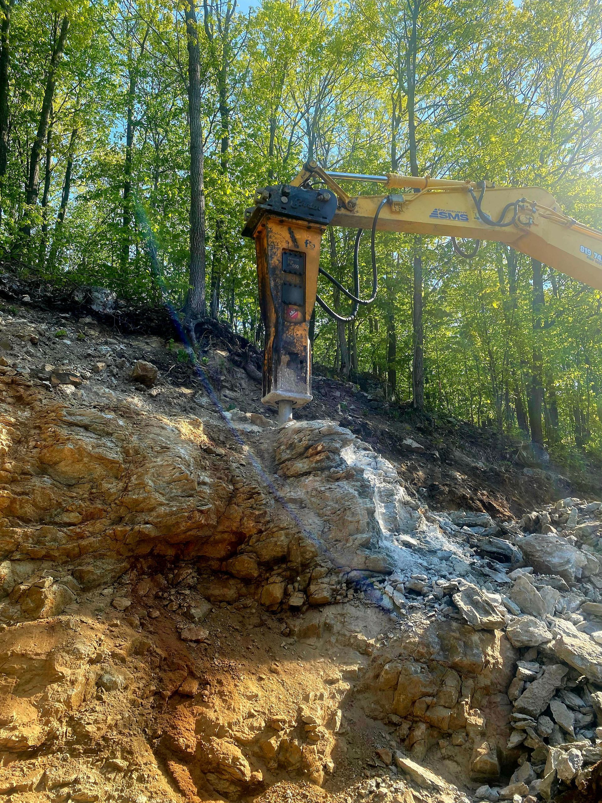 A yellow excavator is working on a rocky hillside in the woods.