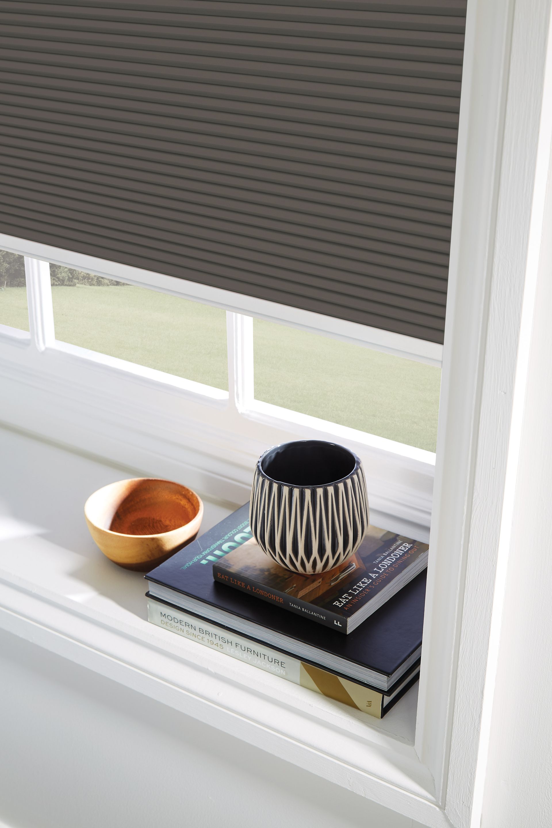 A window with a honeycomb blind on it and a bowl and books on the window sill.