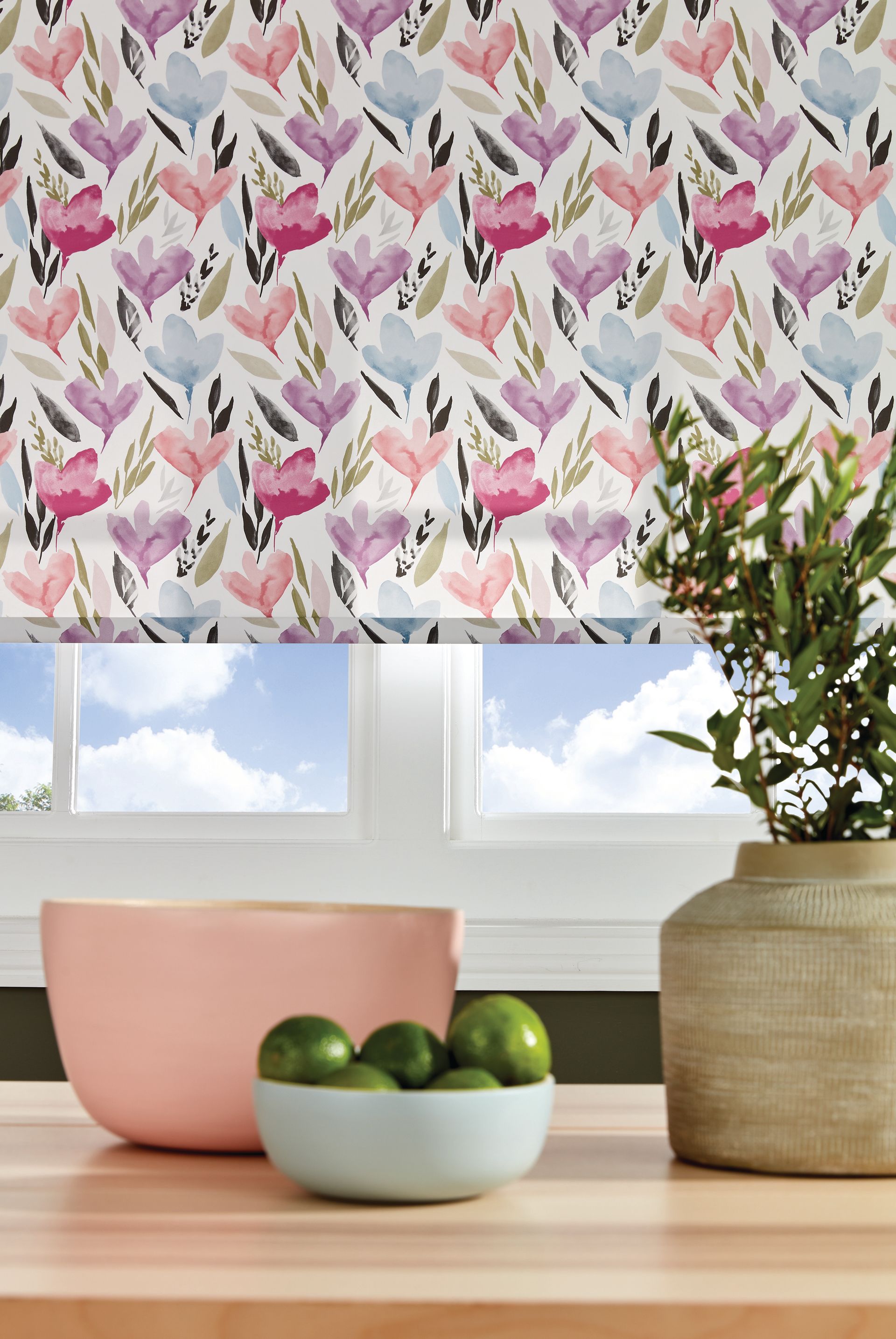 A bowl of limes sits on a wooden table in front of a window.