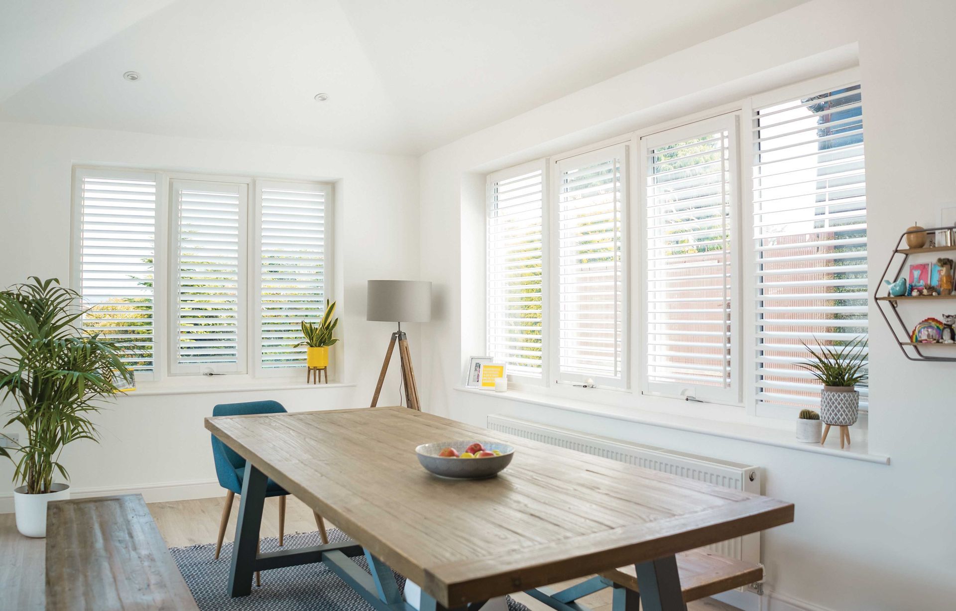 A dining room with a wooden table and chairs and white shutters on the windows.