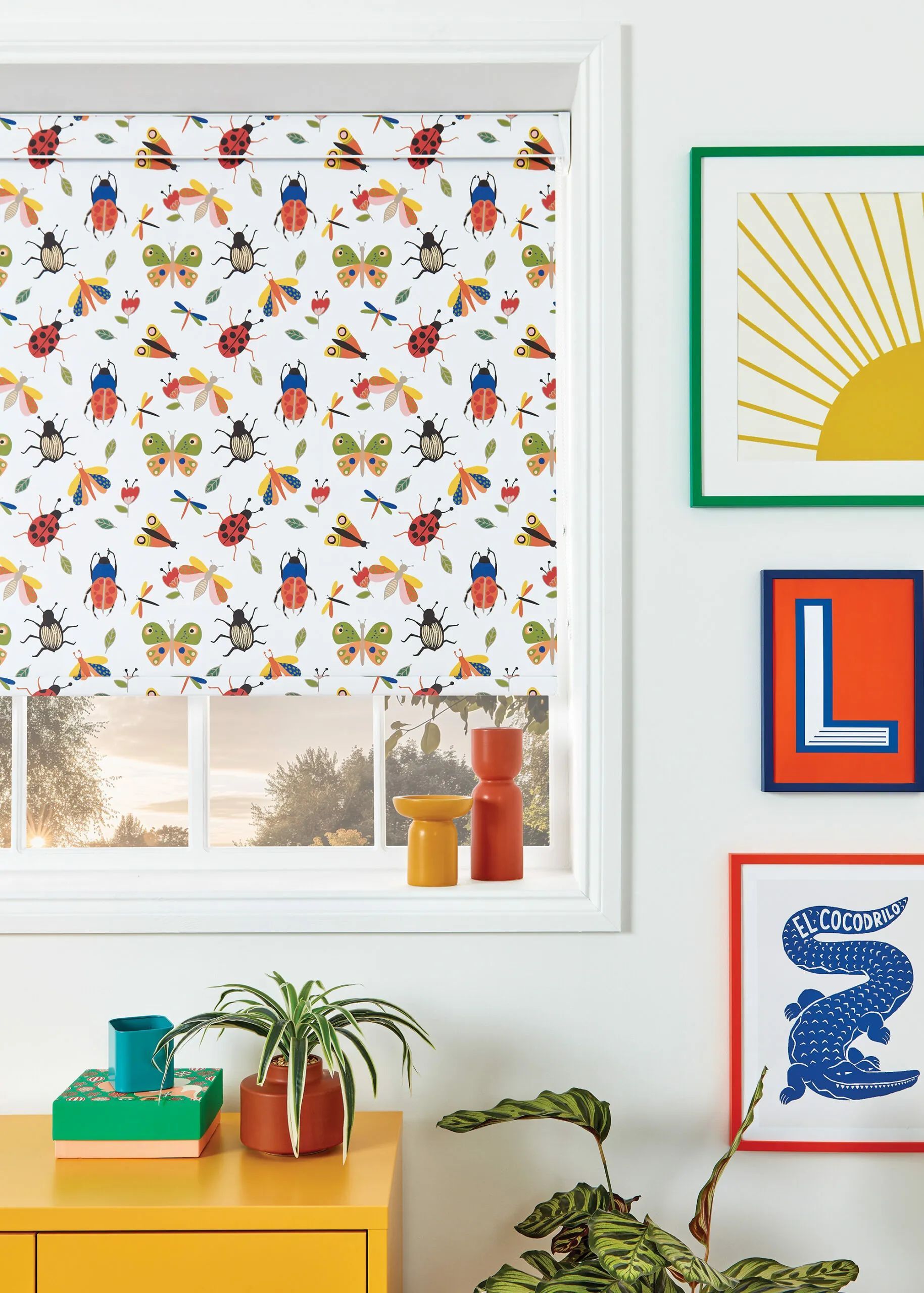 A window with a bug-themed roller blind, colorful framed art, and a yellow table with plants.