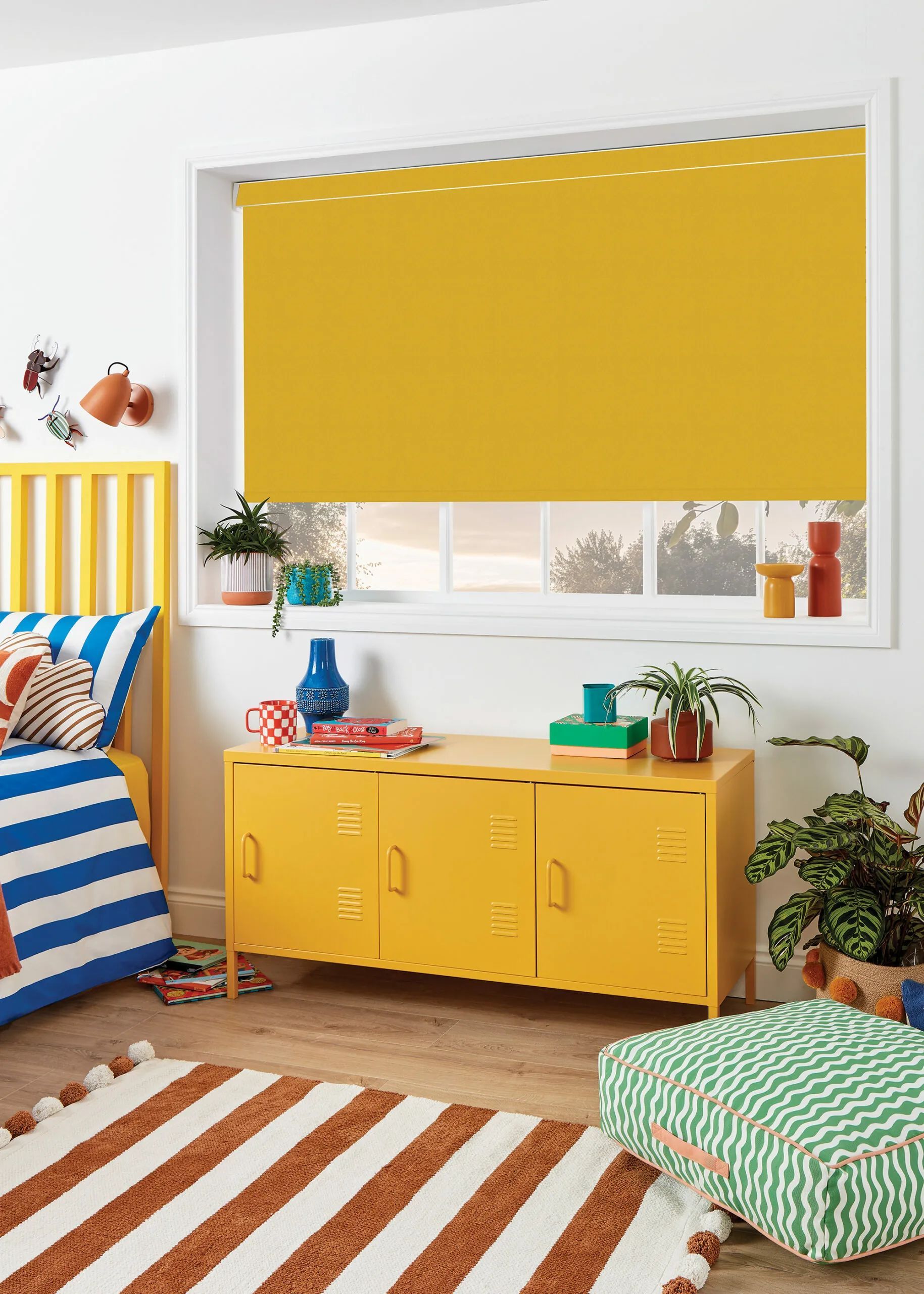 Yellow bedroom with matching blind and cabinet, striped rug, blue bed, and plants.