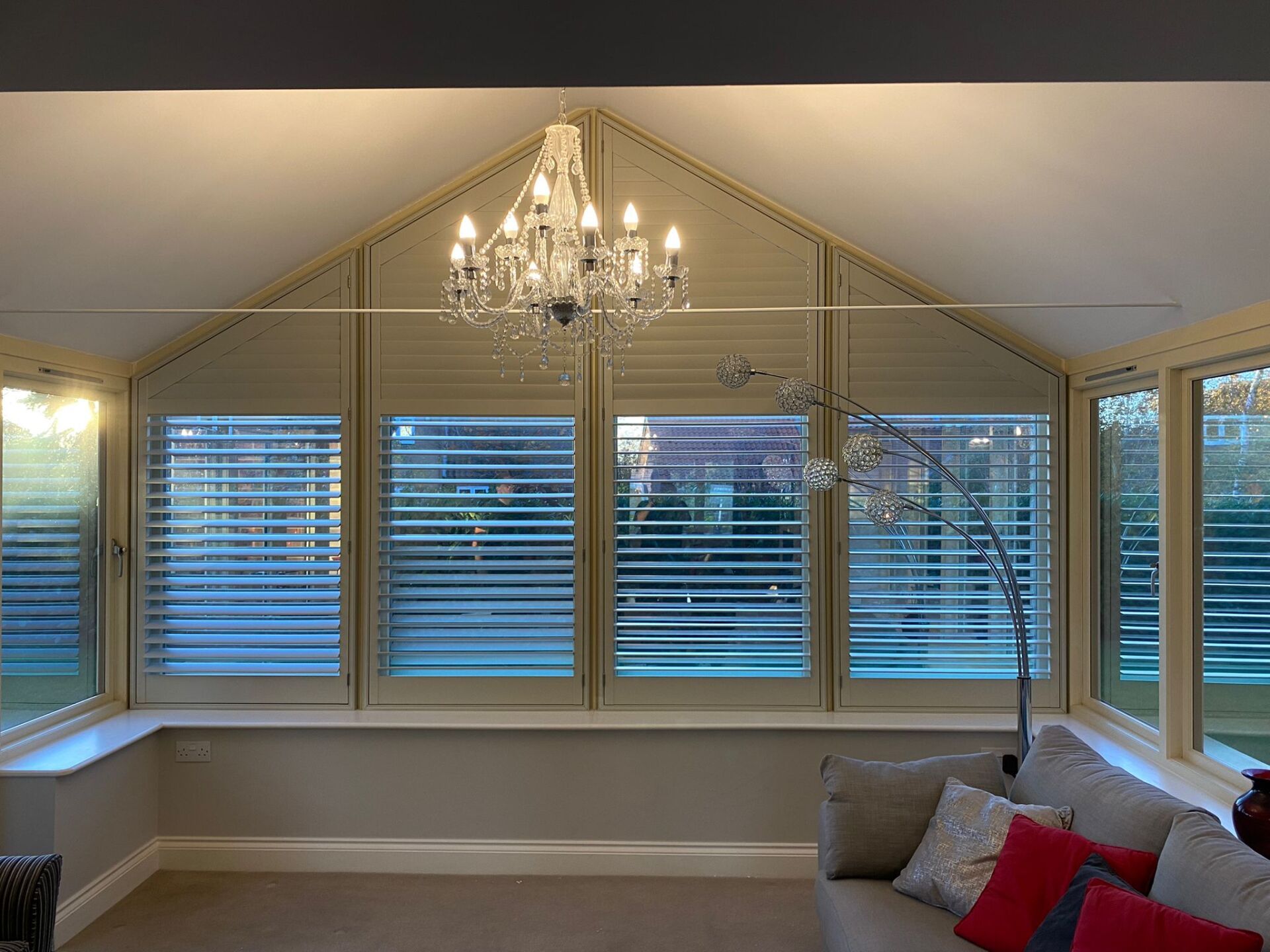 A living room with a couch , chandelier and shutters on the windows.