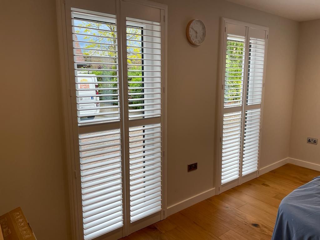 A bedroom with white shutters on the windows and a clock on the wall.