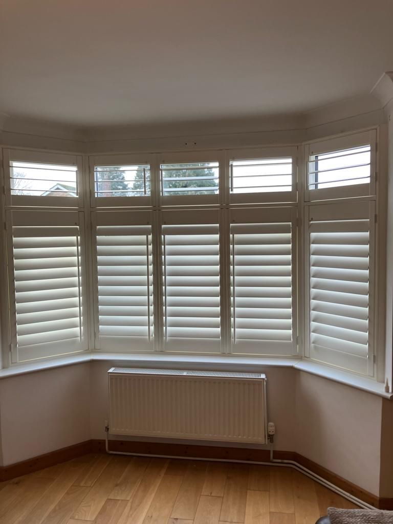 A living room with a large bay window with white shutters and a radiator.