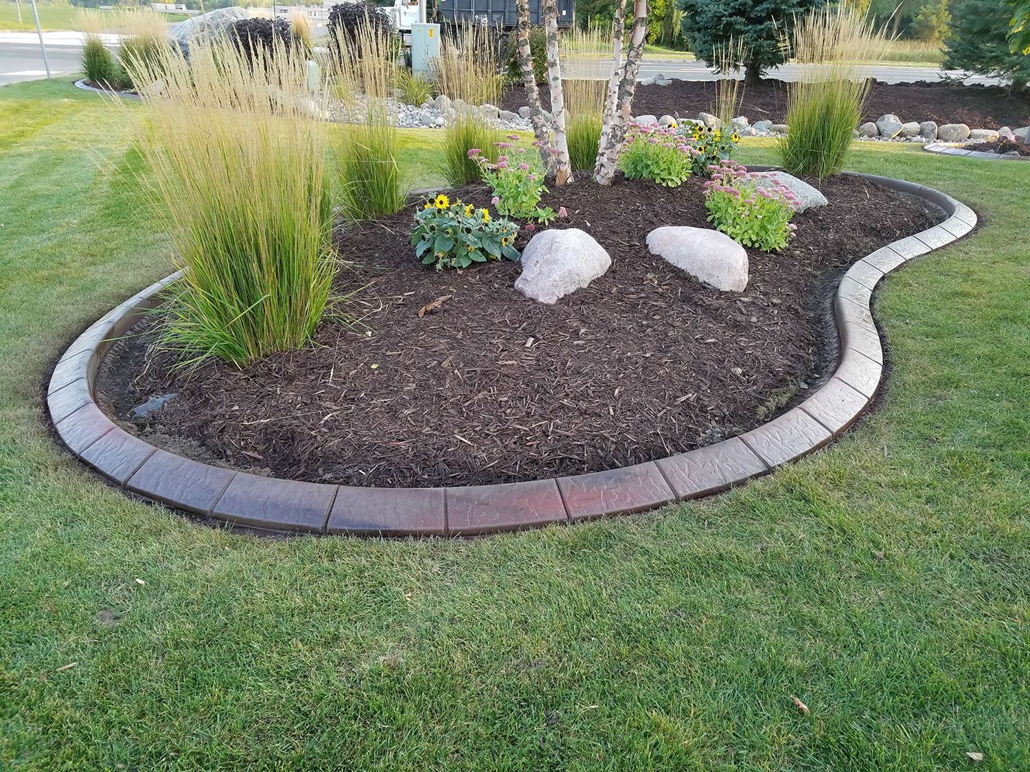 Circular garden bed edged with bricks, containing plants and rocks, in green lawn.