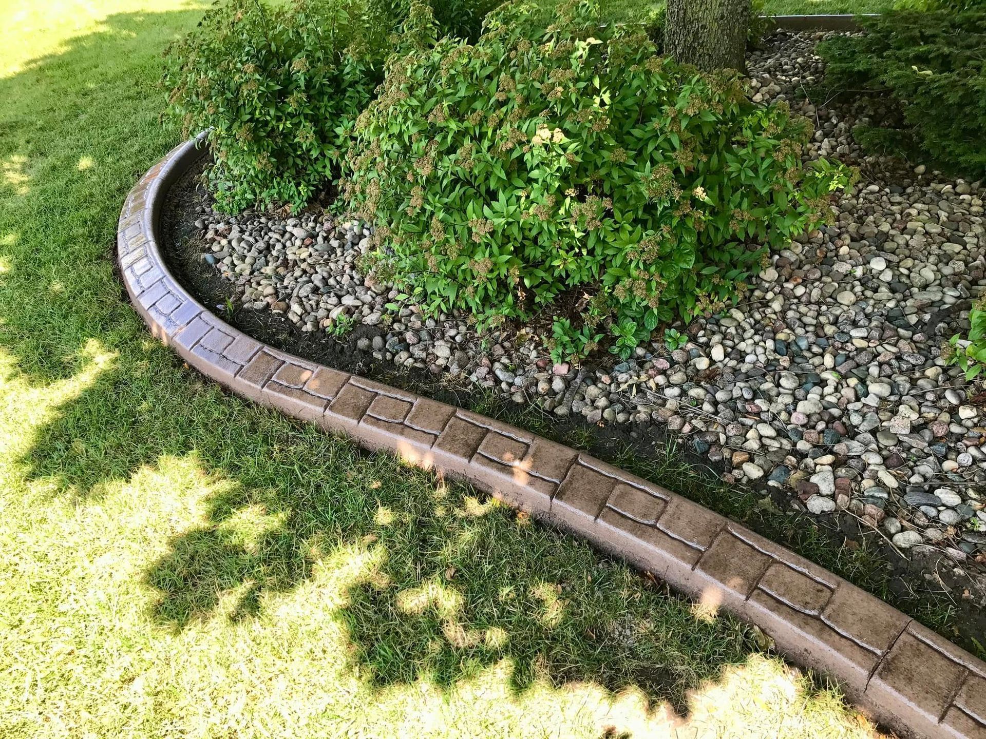 Curved concrete border edging garden bed with green shrubs and gravel, on grassy lawn.