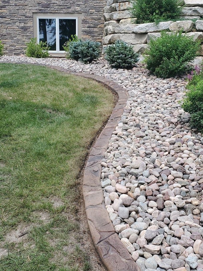 Lawn edged by a concrete border and rocks, next to a building with a window and landscaping.