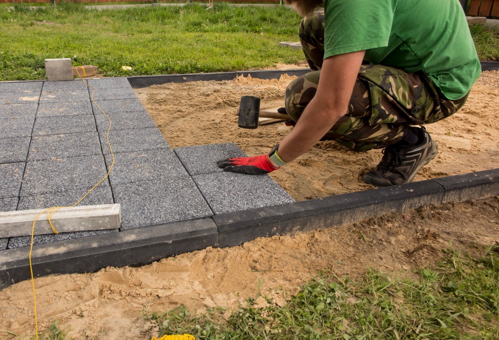 Person laying dark gray paving stones in a sandy area, using a hammer and wearing work gloves.