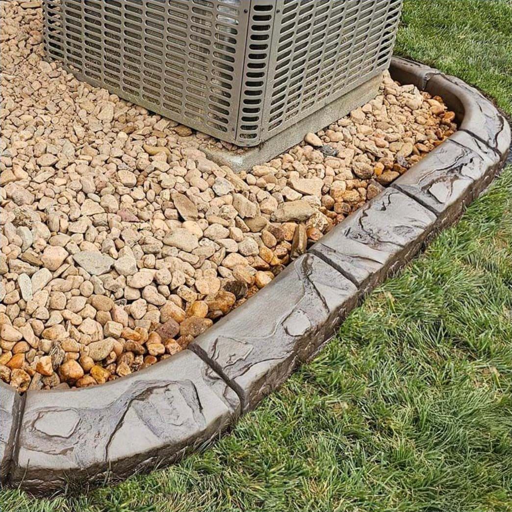 Stone-like concrete edging around an air conditioner unit on gravel, grass visible.