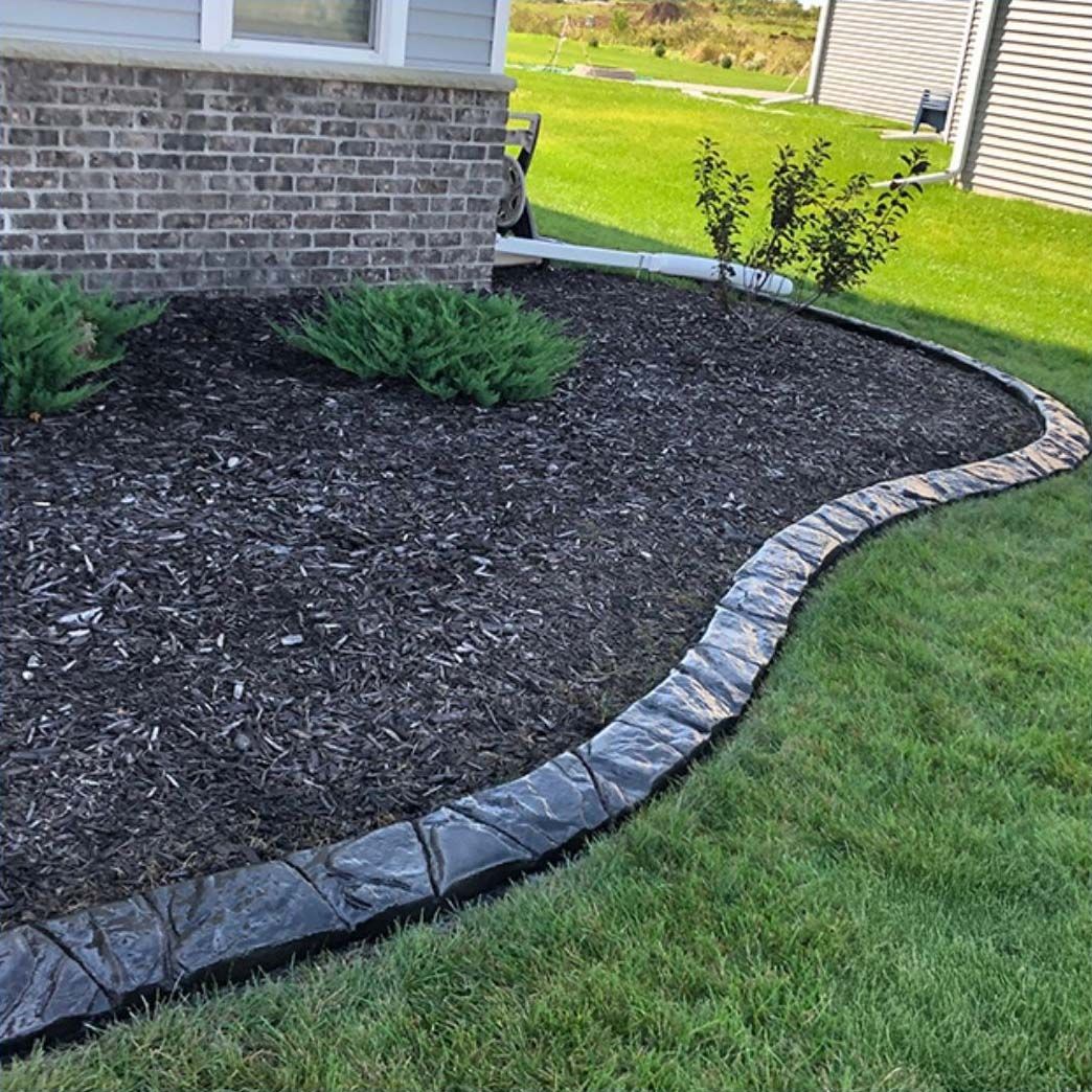 A landscaped garden bed with dark mulch, green shrubs, and a gray stone border, next to green grass and a brick house.