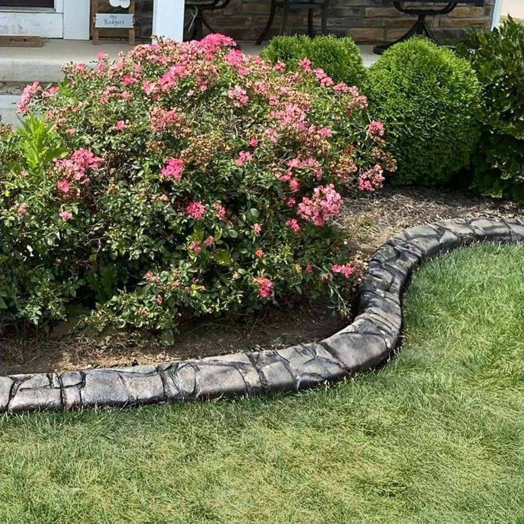 Stone-edged flower bed with pink flowering shrub and green, rounded bushes, set on a green lawn.
