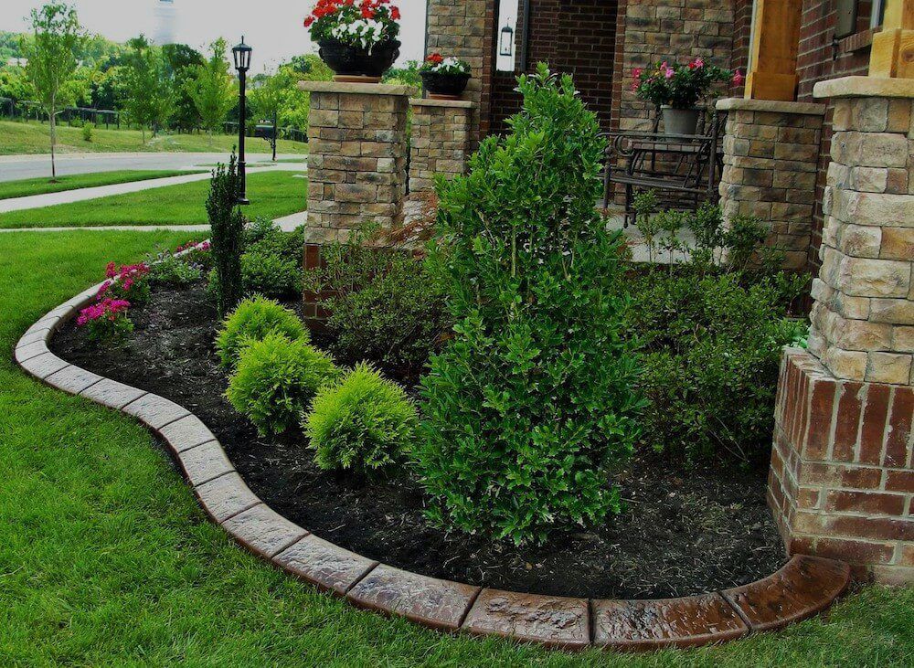 Landscaped garden bed with decorative stone border, green plants, and brick pillars.