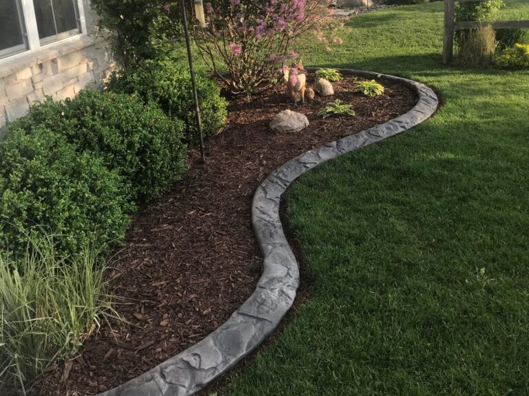 A gray stone-like border curves around a garden bed filled with plants, contrasting with the green grass.