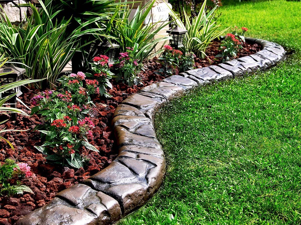 Stone-like concrete edging separates a flower bed with red flowers and greenery from green grass.