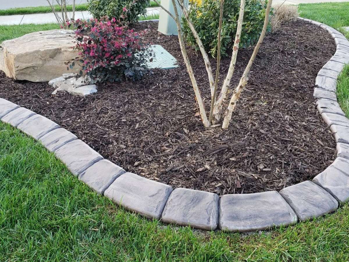 Stone-edged flower bed with brown mulch, plants, and a large rock on green grass.
