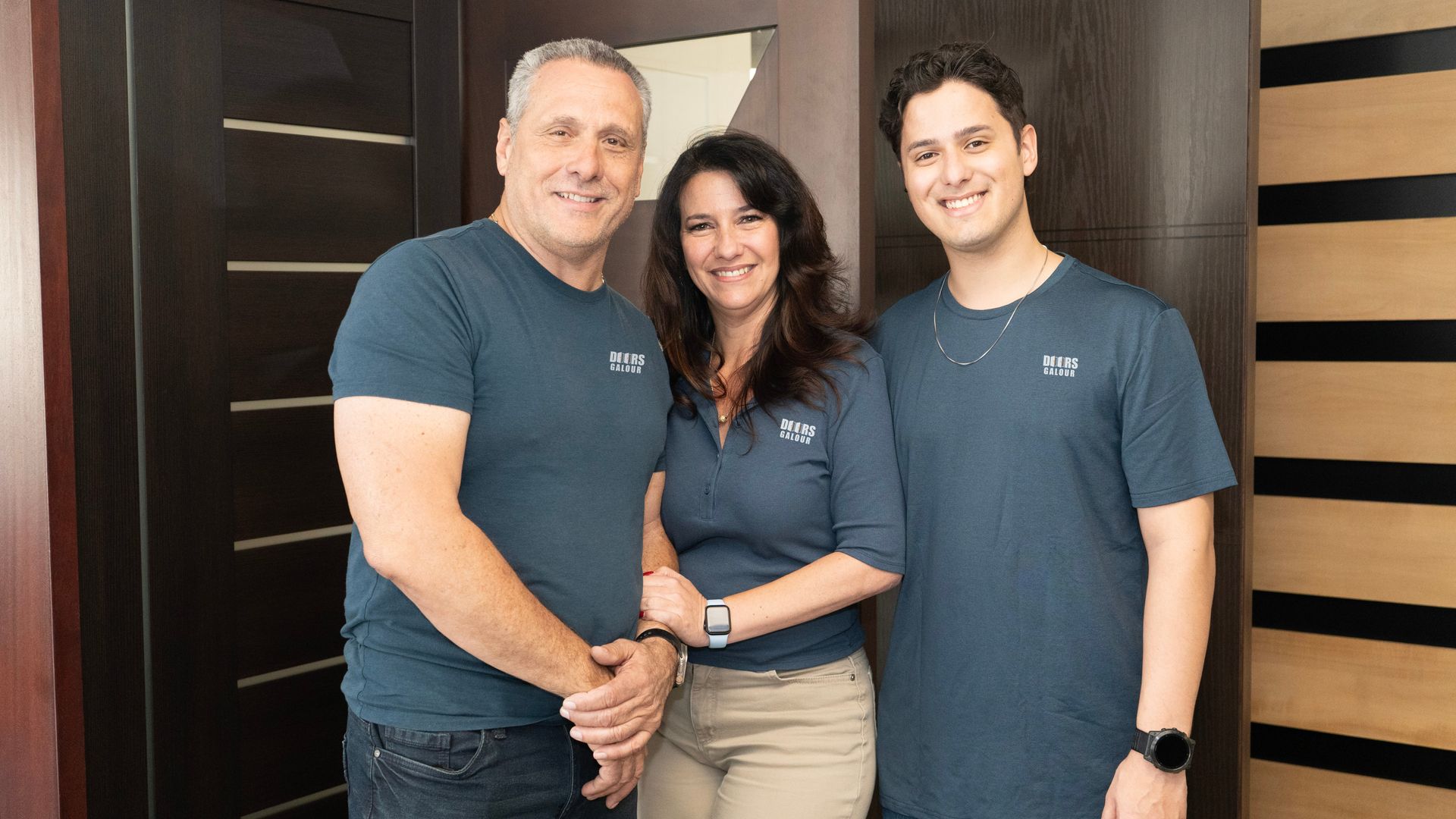 Three people, two men and a woman, standing together in front of a wooden door, all wearing blue shirts, smiling.