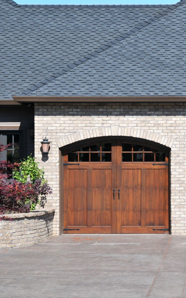 Wooden arched double garage doors on a brick driveway, framed by a stone building, with two sconce lights.