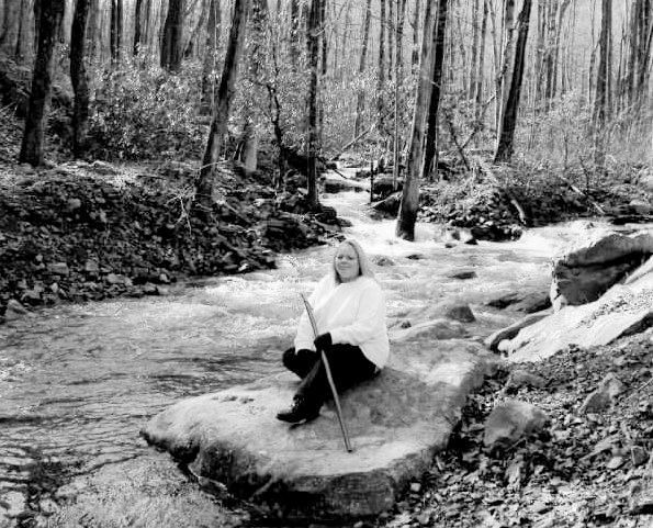 A woman is sitting on a rock next to a stream in the woods.