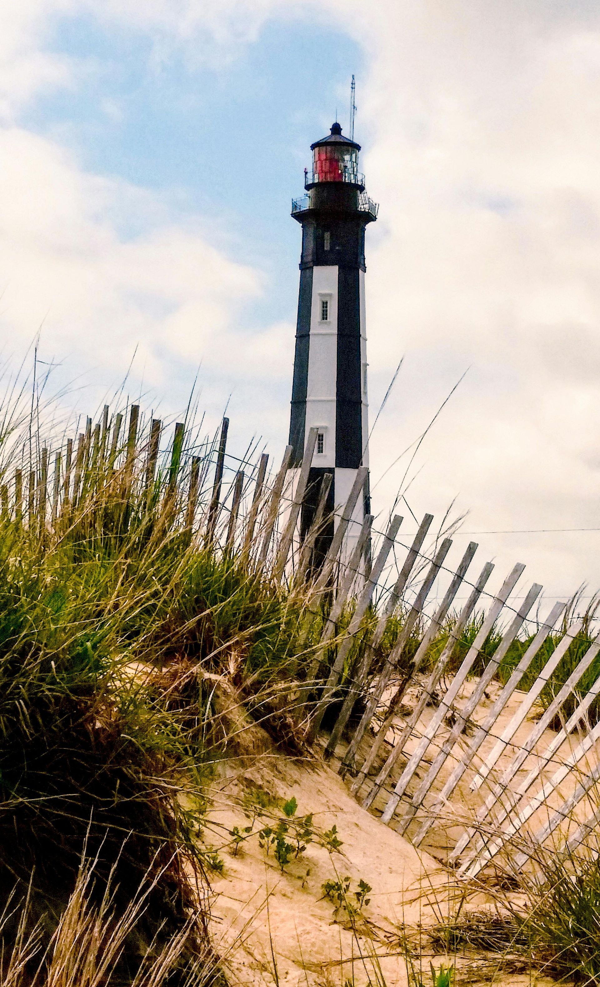 A lighthouse is sitting on top of a sand dune next to a fence.