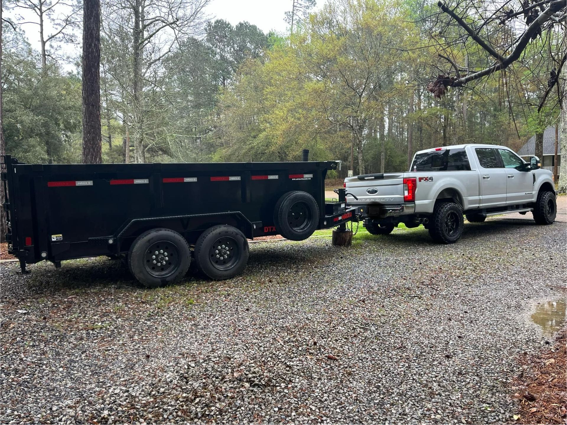 A white Ford pickup truck parked on a gravel driveway, hitched to a large, black dump trailer.