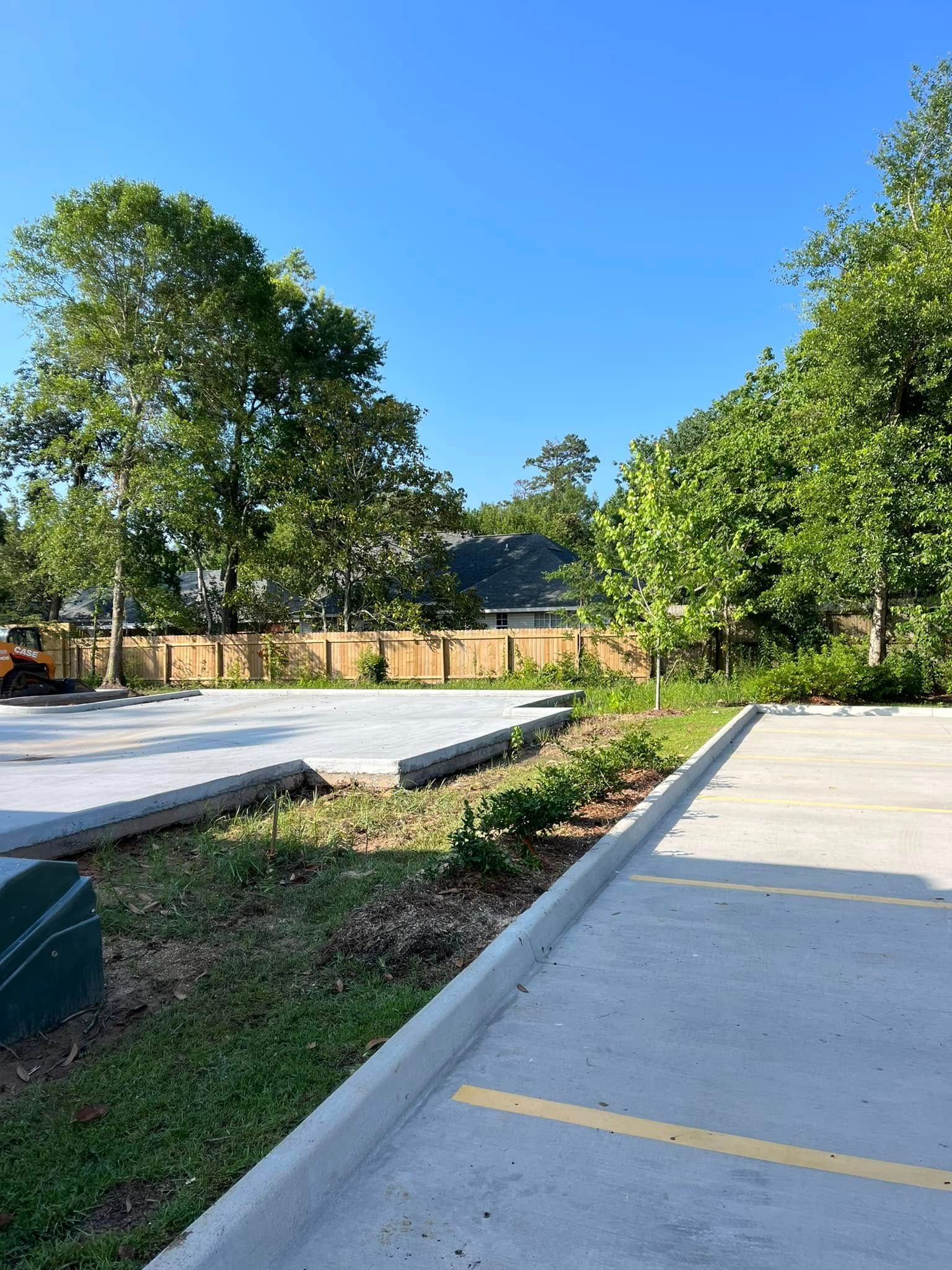 A concrete parking lot area next to a landscaped garden bed with small plants and a tall wooden fence under a clear sky.