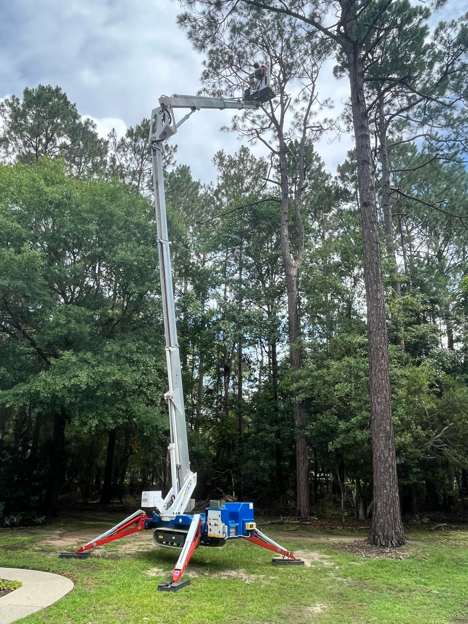 A white aerial lift with extended red outriggers parked on a grassy lawn with trees in the background.