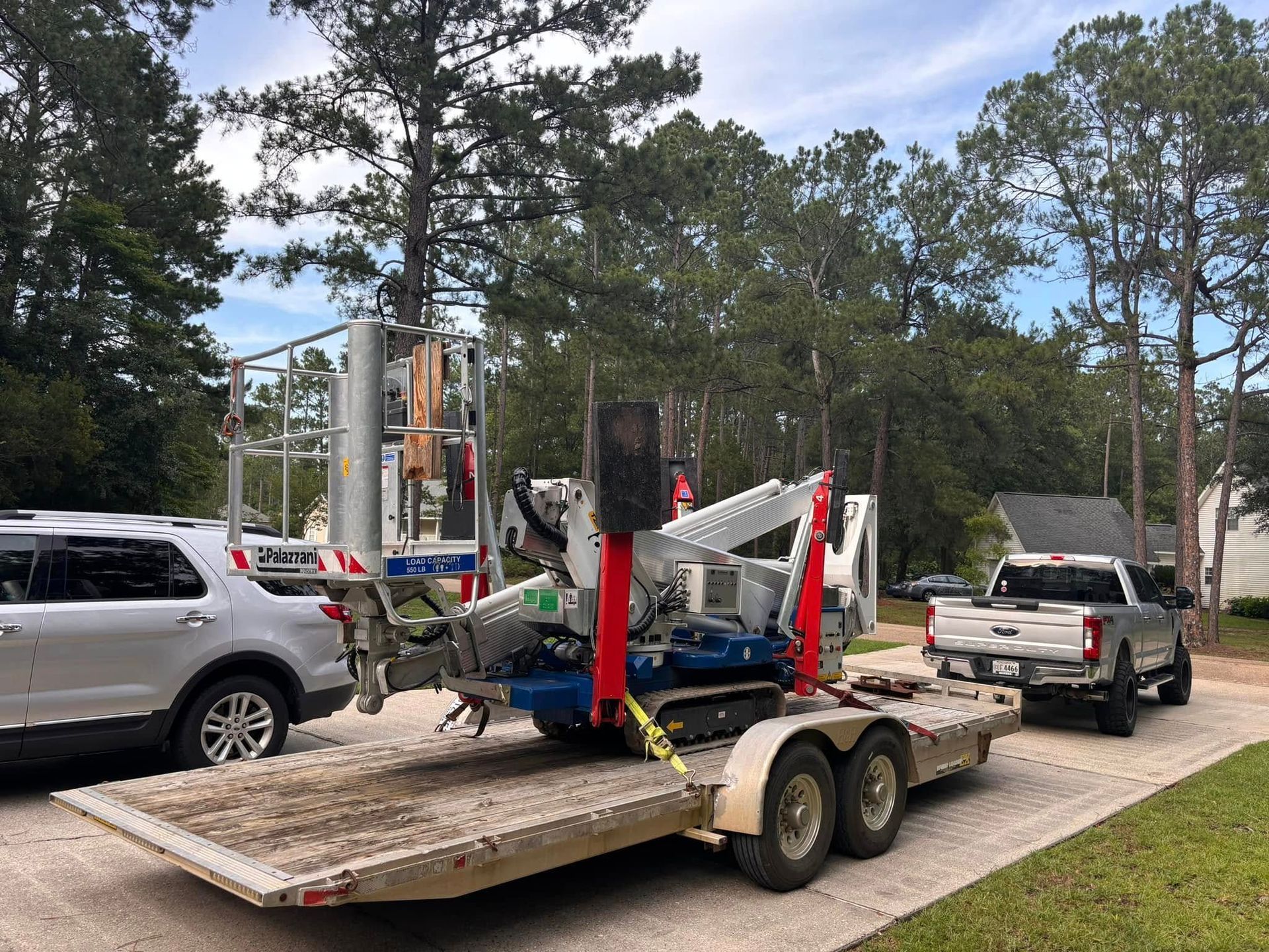 A tracked aerial lift is secured to a flatbed trailer, which is attached to a pickup truck parked on a gravel driveway.