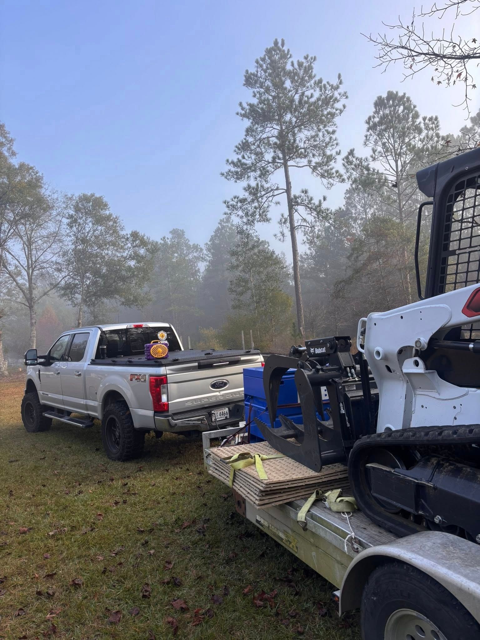 A white pickup truck hauls a trailer carrying a white skid steer with heavy-duty attachments in a foggy, wooded area.