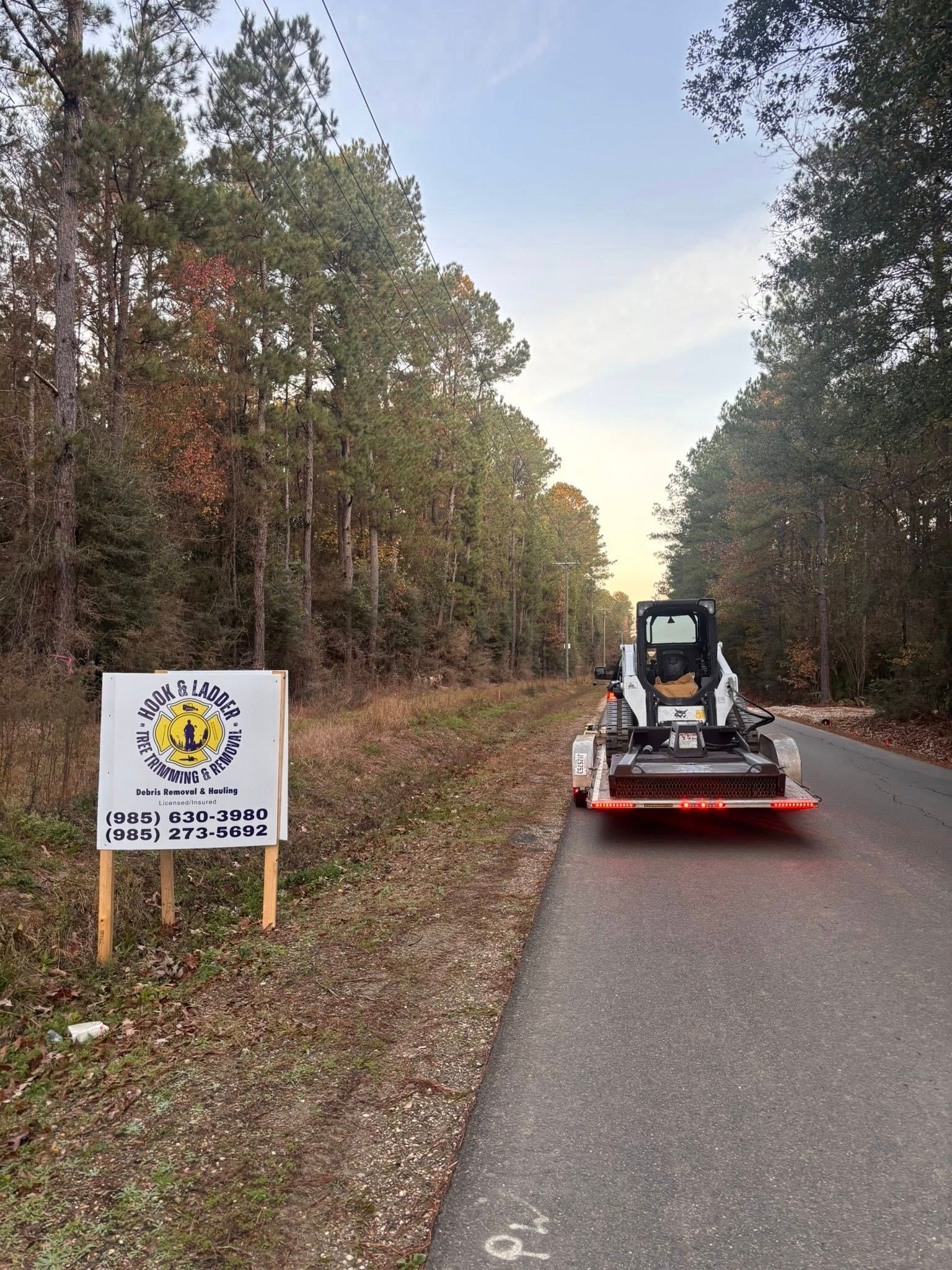 A skid steer loader on a trailer parked on a rural road next to a sign for a forestry mulching service.