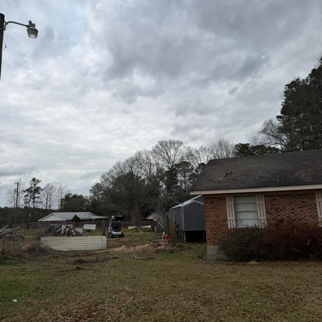 A brick house on the right with a yard containing a shed, a small tractor, and scattered debris under a cloudy sky.