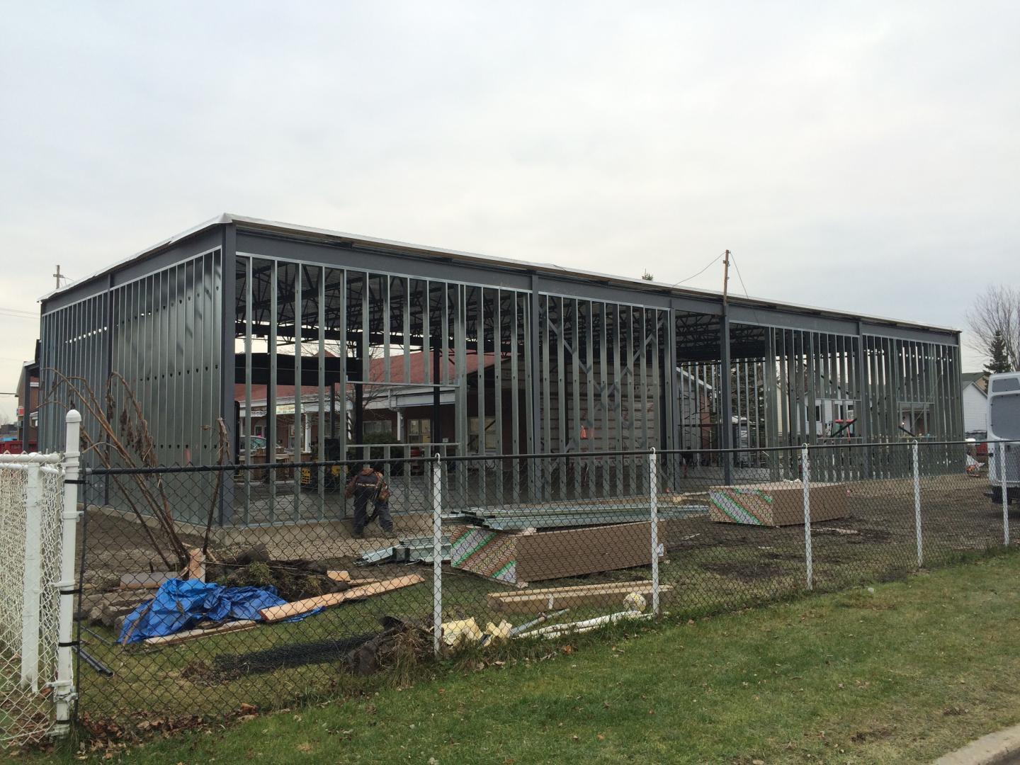 Un grand bâtiment métallique est en cours de construction à côté d'une clôture à mailles losangées.