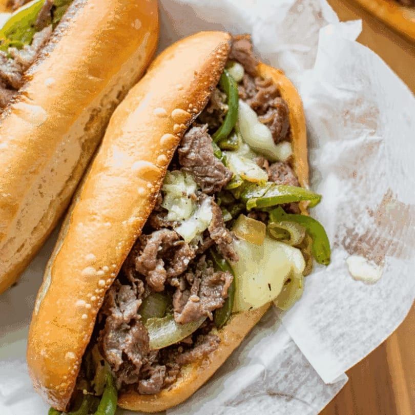 Two cheesesteak sandwiches on white paper, with toasted rolls, beef, onions, and green peppers.