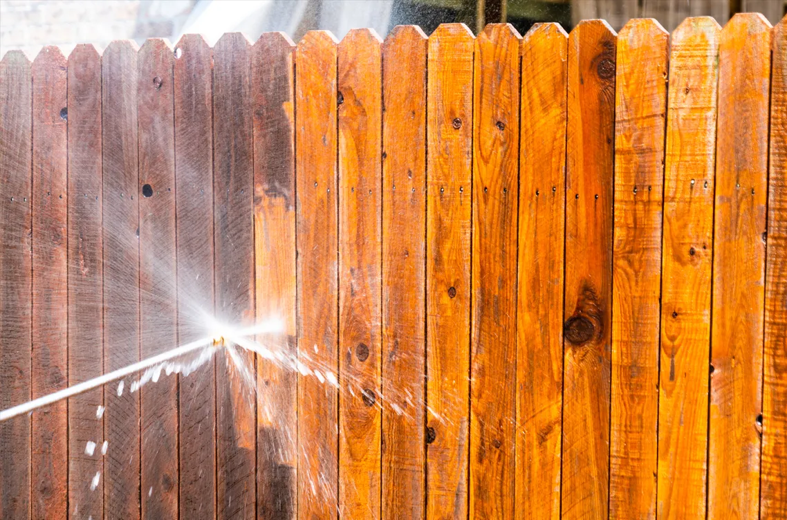 A person is cleaning a wooden fence with a high pressure washer.