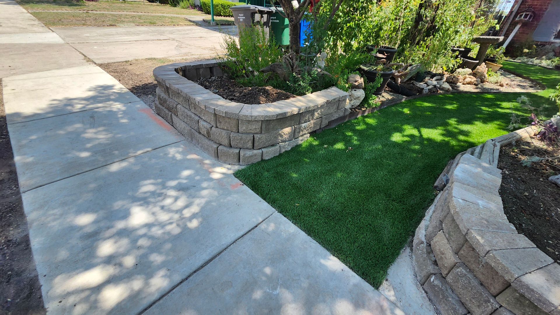 Concrete sidewalk curves past a low retaining wall with grass and a flower bed.