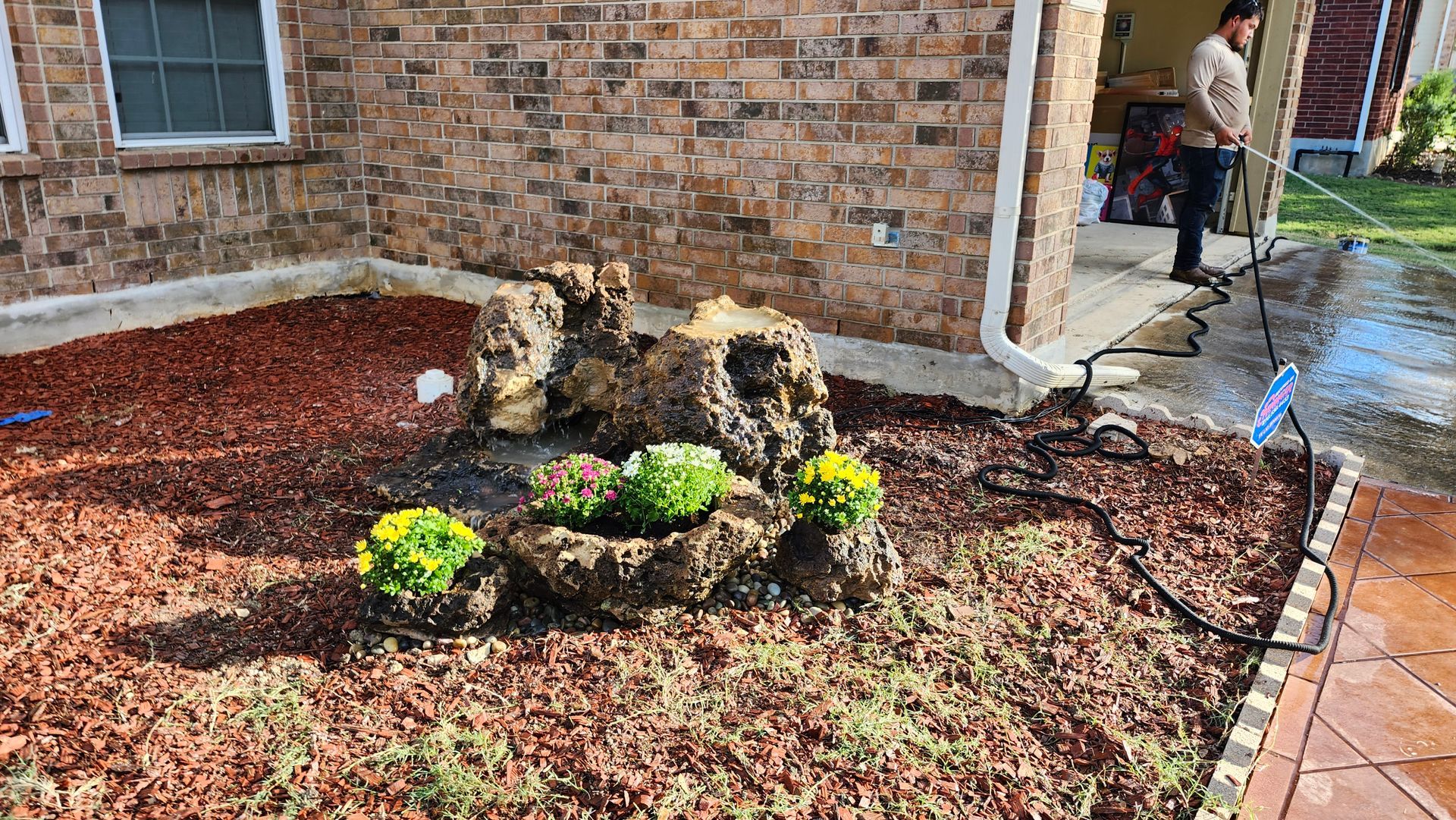 Person pressure washing a yard near a rock and flower garden with brick house in the background.