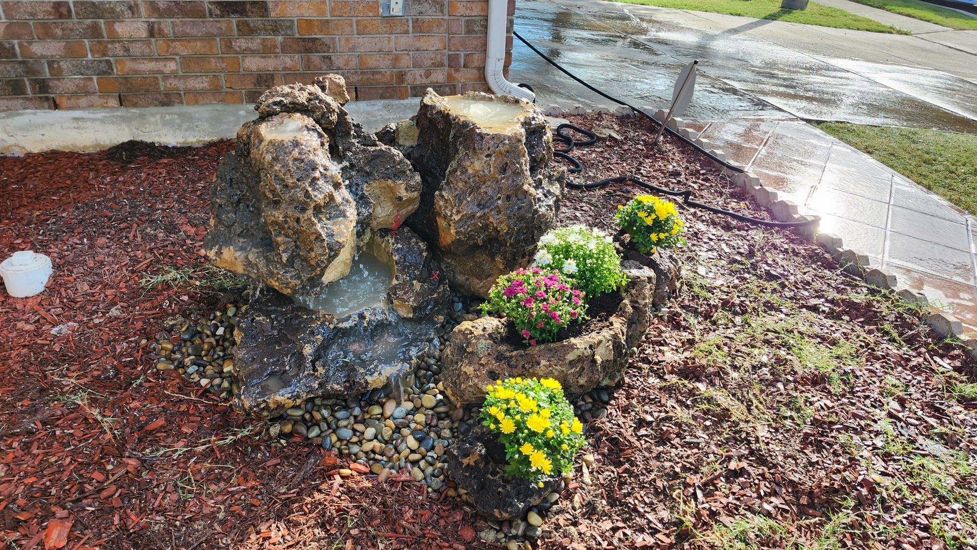Rock water feature with colorful flowers, set in a mulch bed, next to a sidewalk and a building.