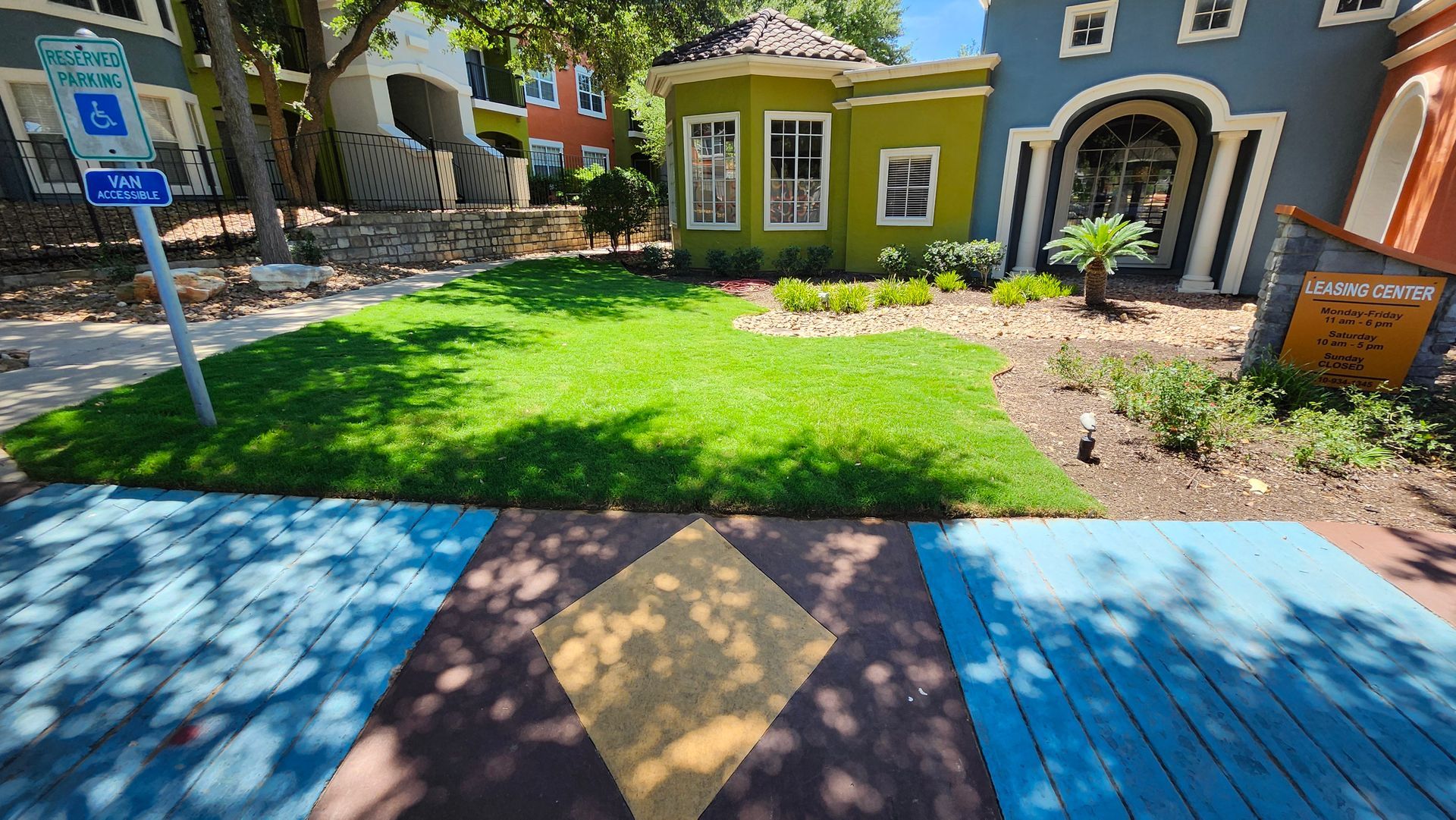 Street with colorful buildings, green lawn, blue and brown painted sidewalk, and a handicap parking sign.